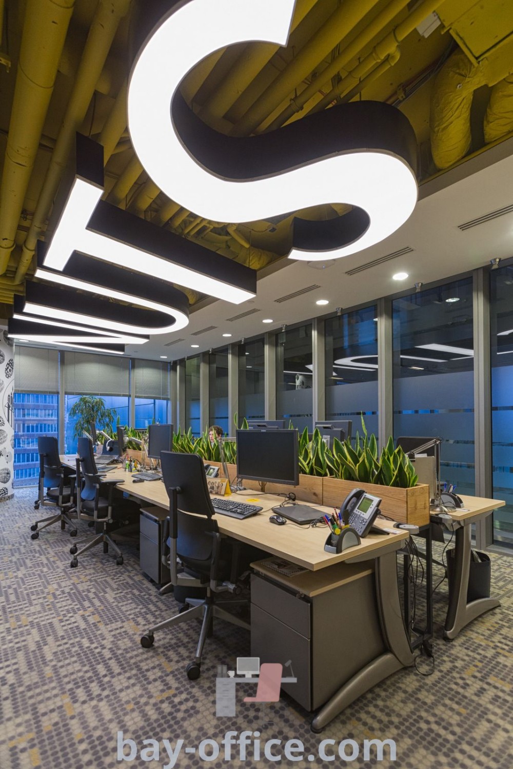 Open office design with two desks, computers, and planters near large windows, showcasing modern and industrial office aesthetics. Explore design trends and decor ideas that inspire productivity at bay-office.com.
