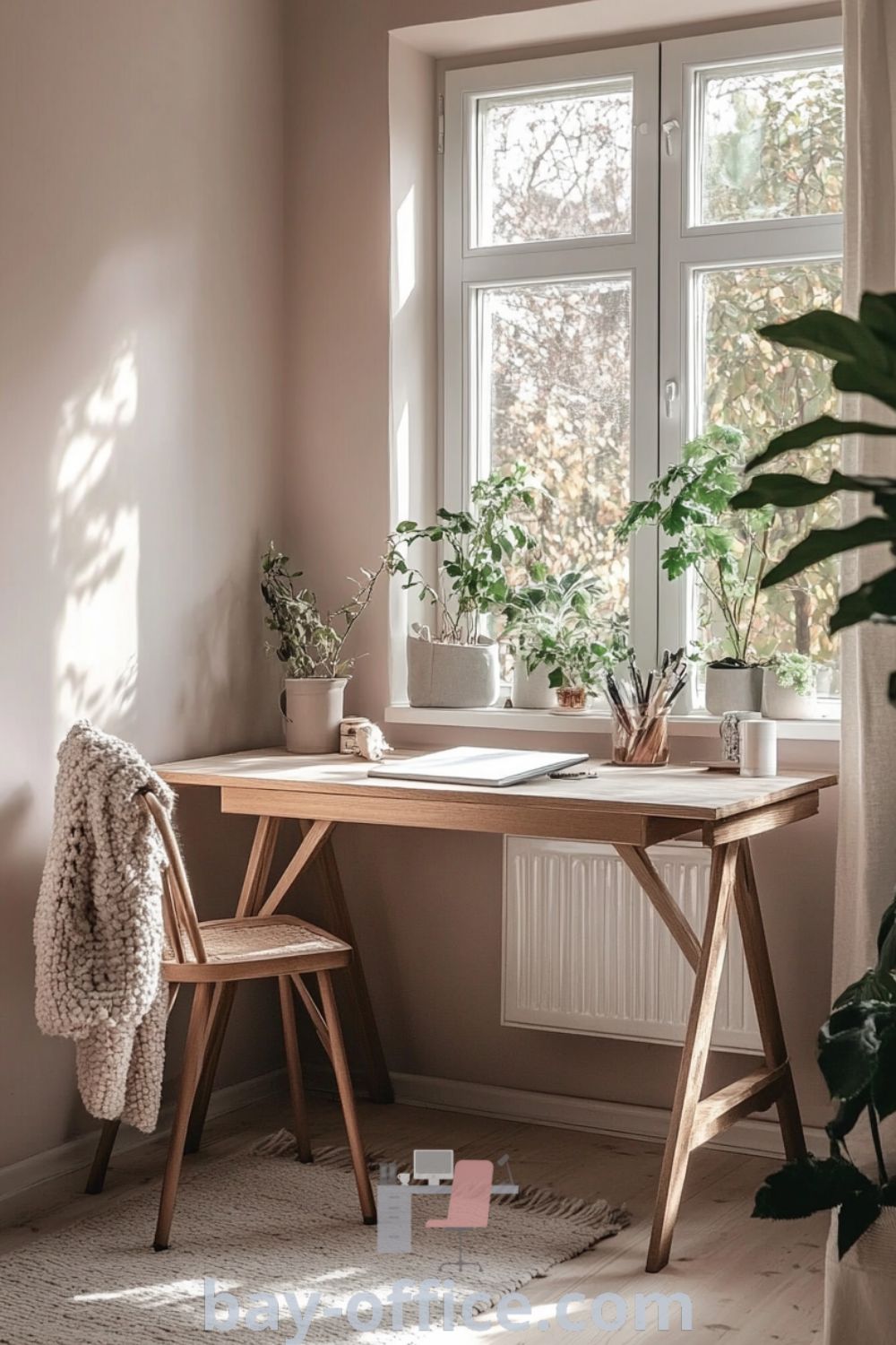 Cozy Scandinavian home office featuring soft pink walls, natural wood furniture, and sunlight streaming through a large window. The design includes minimalist decor, textured throws, and greenery, creating an inspiring workspace that enhances focus and comfort. Explore more design ideas at bay-office.com.